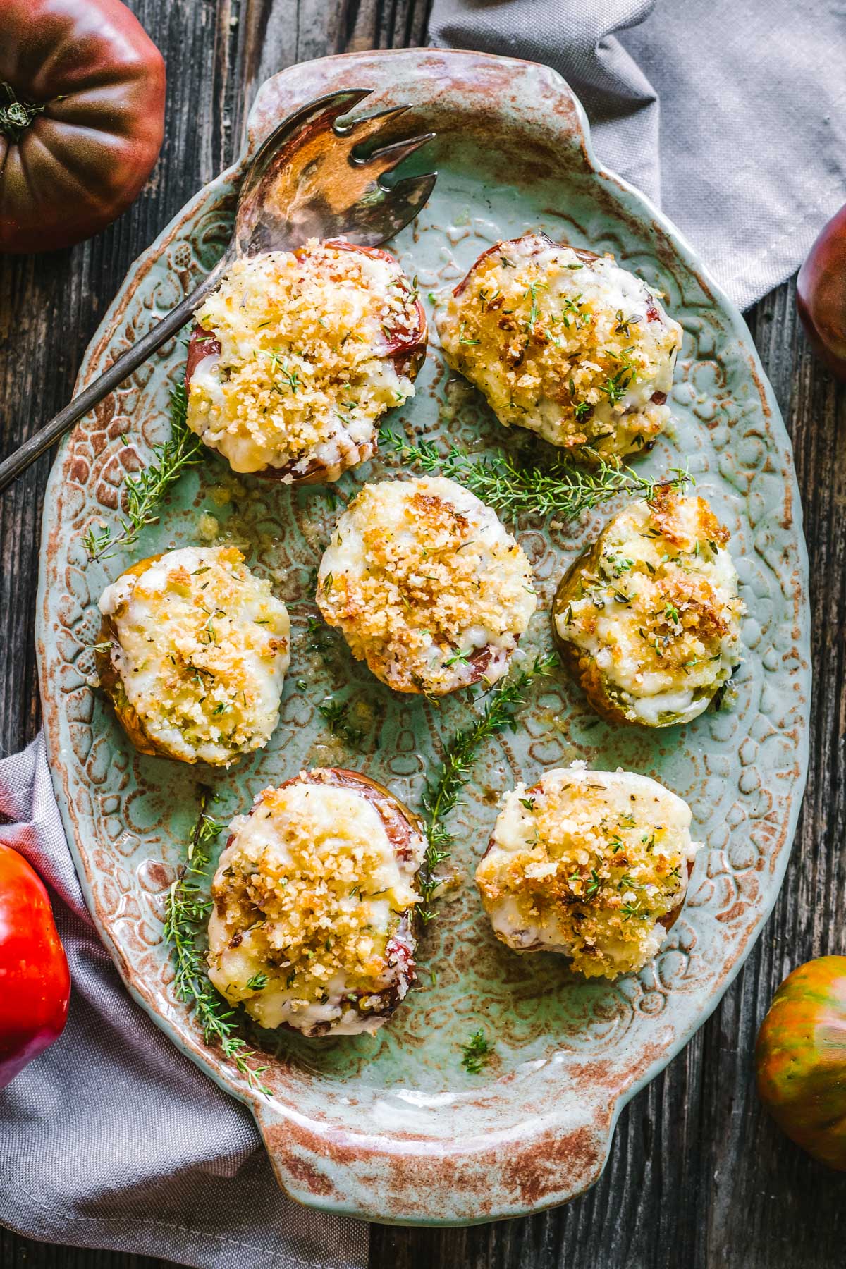 Top-down view of seven baked tomato halves arranged neatly on a textured green platter with sprigs of thyme and a vintage serving spoon.
