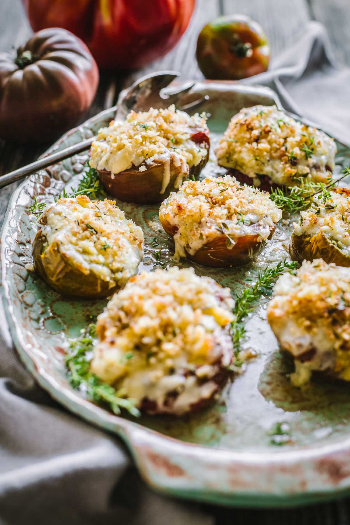 Angled perspective showing baked cheesy tomatoes with a crunchy topping, garnished with herbs and surrounded by heirloom tomatoes.