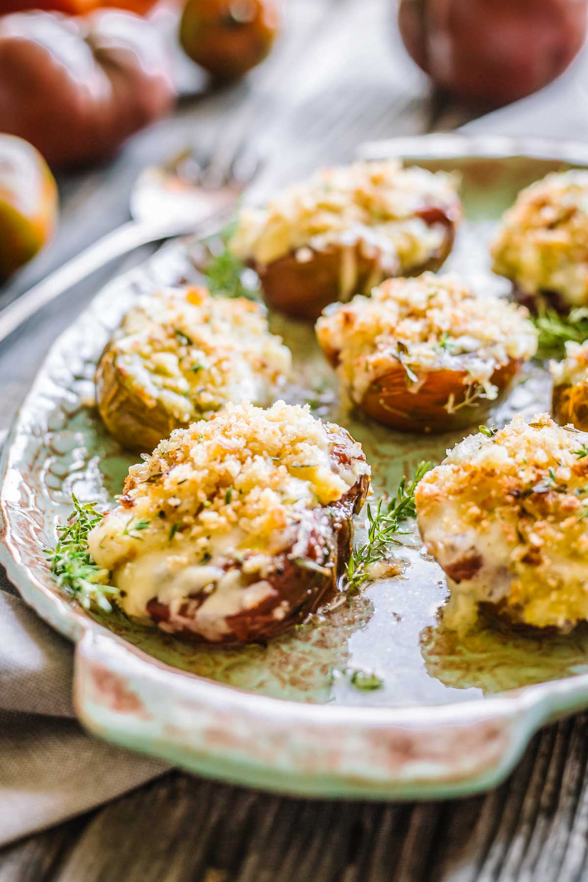 Sharp foreground focus on cheesy, breadcrumb-topped baked tomatoes with melted cheese edges and flecks of green herbs.