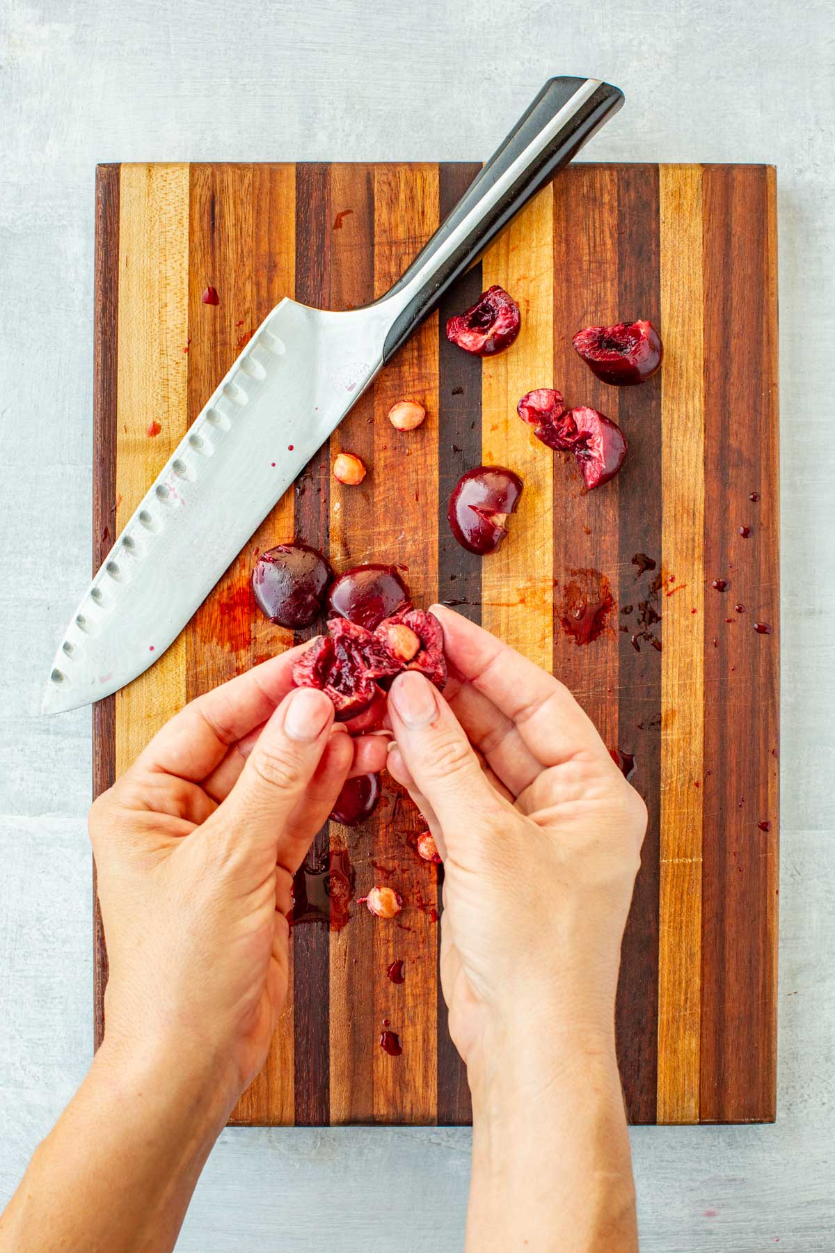 Hands splitting open a cherry to remove the pit on a striped wooden cutting board with a chef's knife and scattered cherry pits.