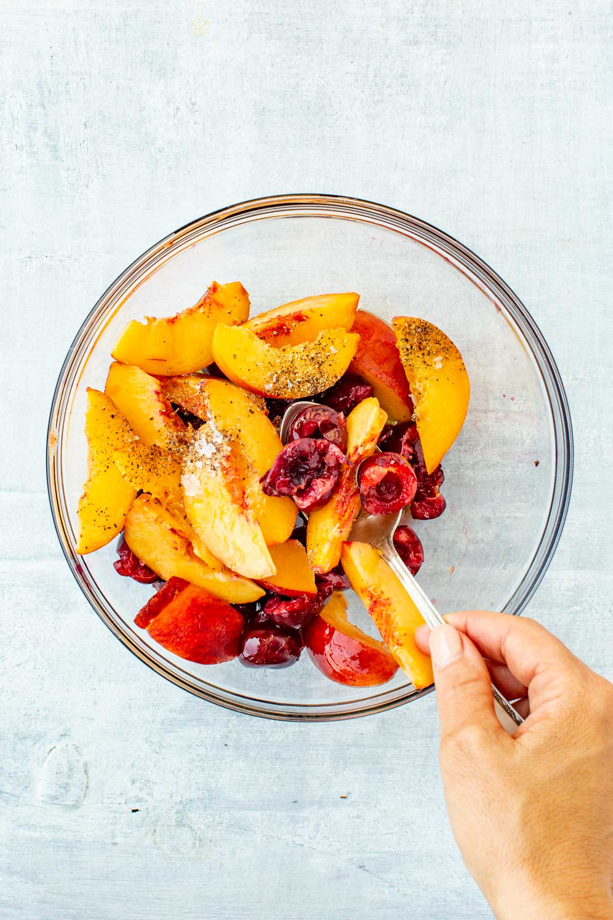 Overhead view of a glass bowl filled with sliced peaches and halved cherries being tossed with salt and pepper.