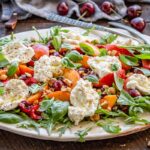 Side angle of the full salad platter showing volume and color with fresh basil, peaches, cherries, and burrata on a rustic wooden table.