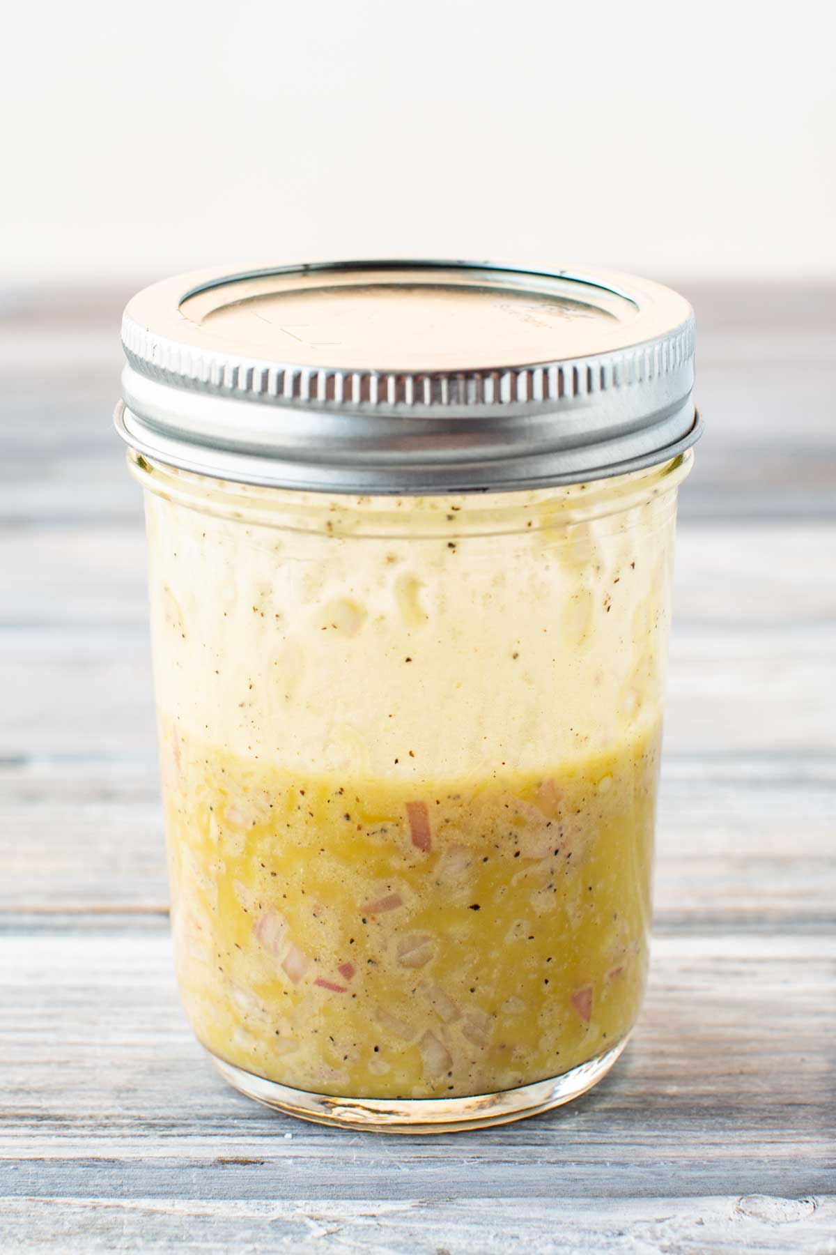 Close-up of a mason jar filled with freshly shaken vinaigrette, showing a creamy, emulsified texture with visible shallots and pepper flecks.
