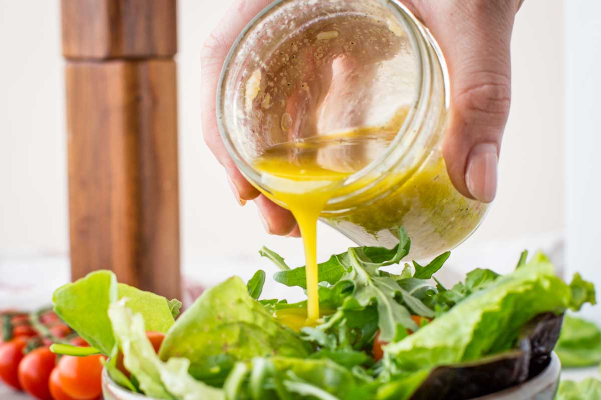 A hand pouring freshly made vinaigrette over a bowl of leafy green salad, with arugula and romaine in the foreground.