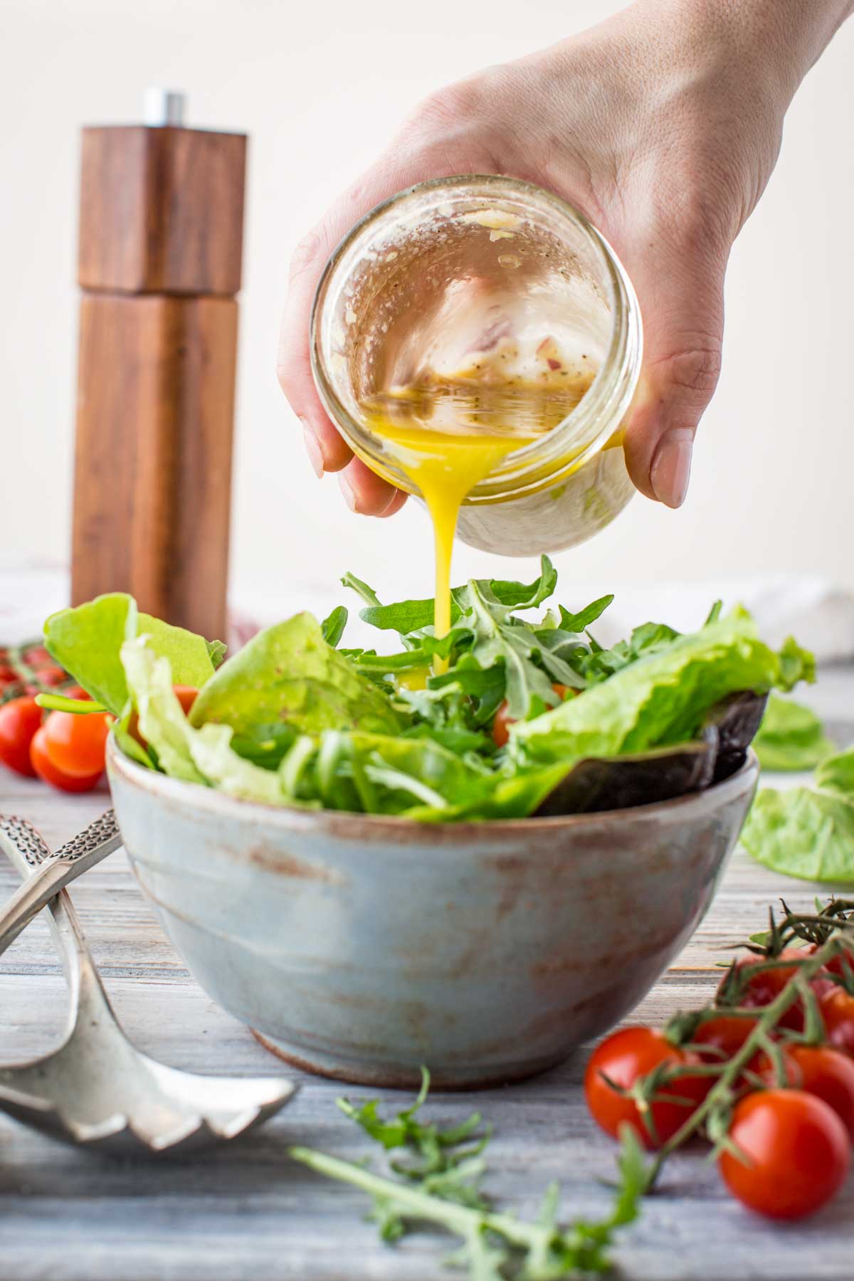 Overhead view of vinaigrette being drizzled onto a green salad in a ceramic bowl, surrounded by cherry tomatoes and salad tongs.