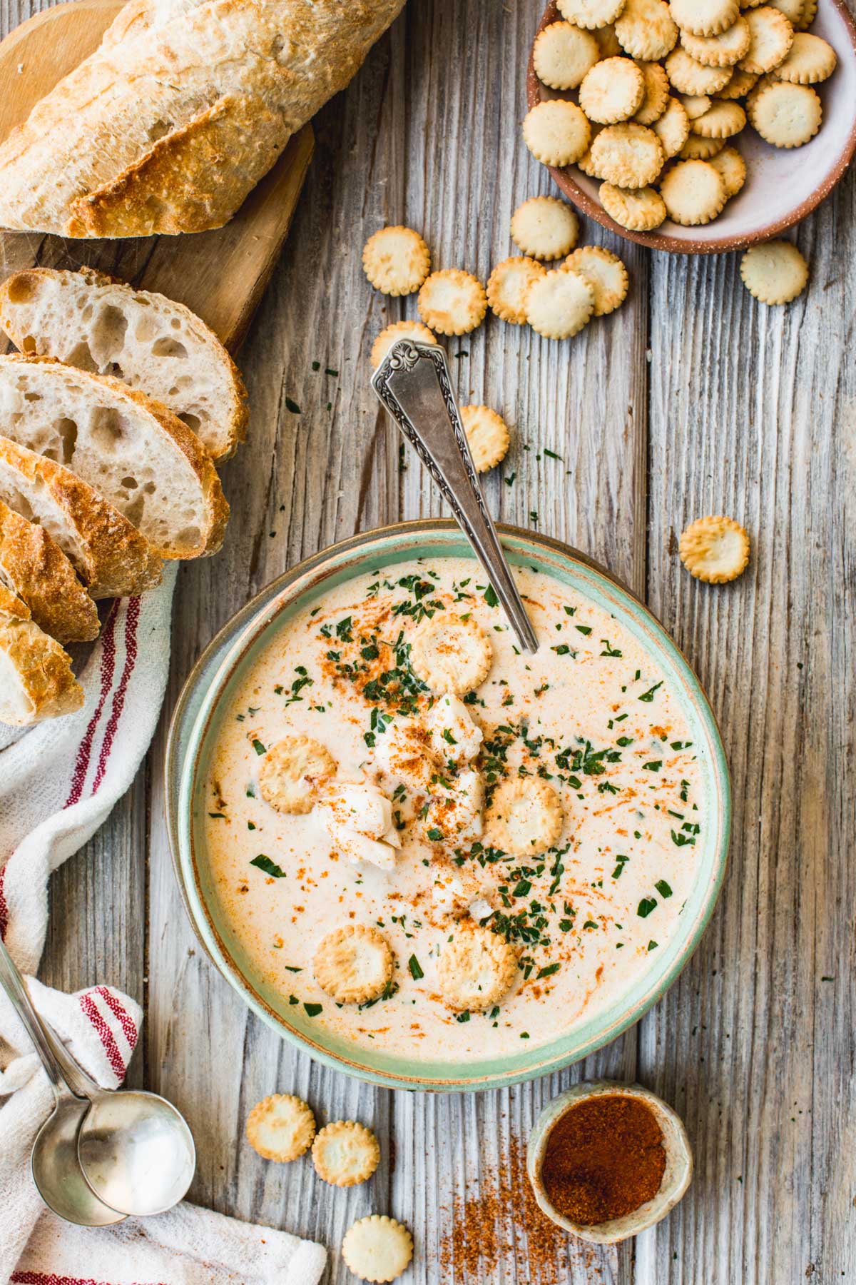 Flat lay of crab soup next to sliced bread, oyster crackers, and a small bowl of Old Bay seasoning on a wooden table.