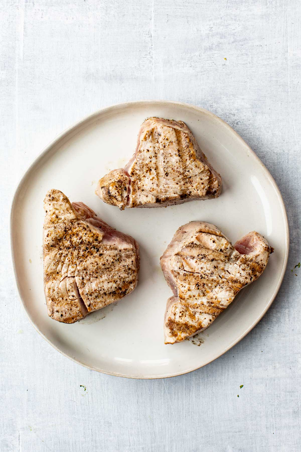 Three grilled tuna steaks on a white ceramic plate, seasoned with salt and pepper and lightly charred on the outside.