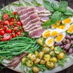 Wide horizontal image of a classic Niçoise salad with all components evenly layered on a bed of arugula and garnished with fresh herbs and flaky sea salt.