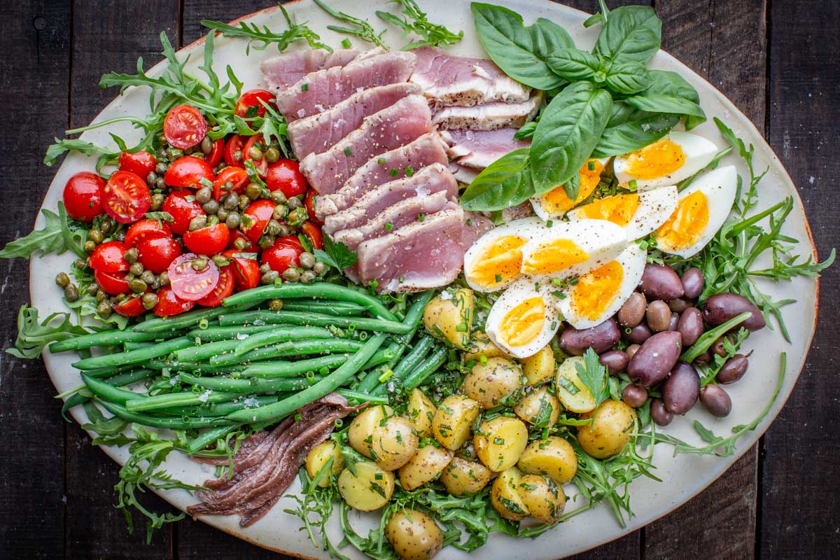 Wide horizontal image of a classic Niçoise salad with all components evenly layered on a bed of arugula and garnished with fresh herbs and flaky sea salt.