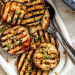 A close-up of grilled eggplant slices on a white oval platter, garnished with chopped herbs and garlic from the marinade.