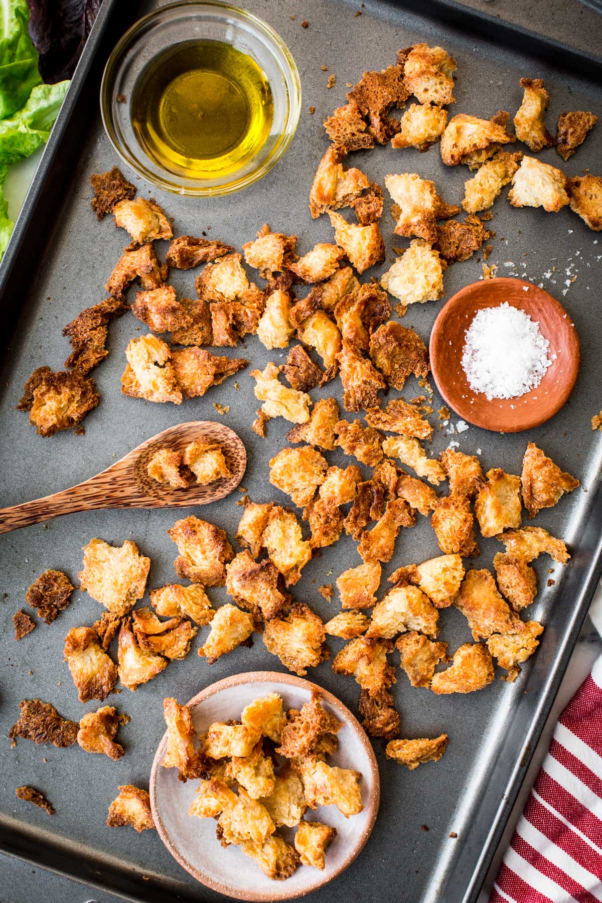 Flat lay of croutons on a tray with bowls of salt and oil, a wooden spoon, and greens in the corner.
