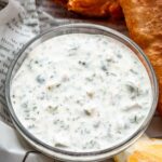 A small glass bowl filled with creamy tartar sauce, speckled with chopped herbs and pickles, placed next to crispy golden fried fish on newspaper-lined parchment.