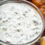A close-up of thick tartar sauce in a bowl, showing finely chopped herbs, capers, and pickles suspended in a smooth, creamy base. Golden fried fish is partially visible in the background.