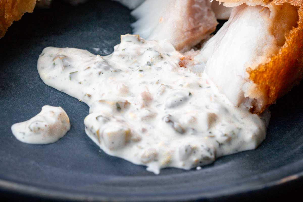 A close-up of tartar sauce spread onto a plate next to a piece of flaky, battered fried fish. The sauce has a chunky texture with visible herbs and pickles.
