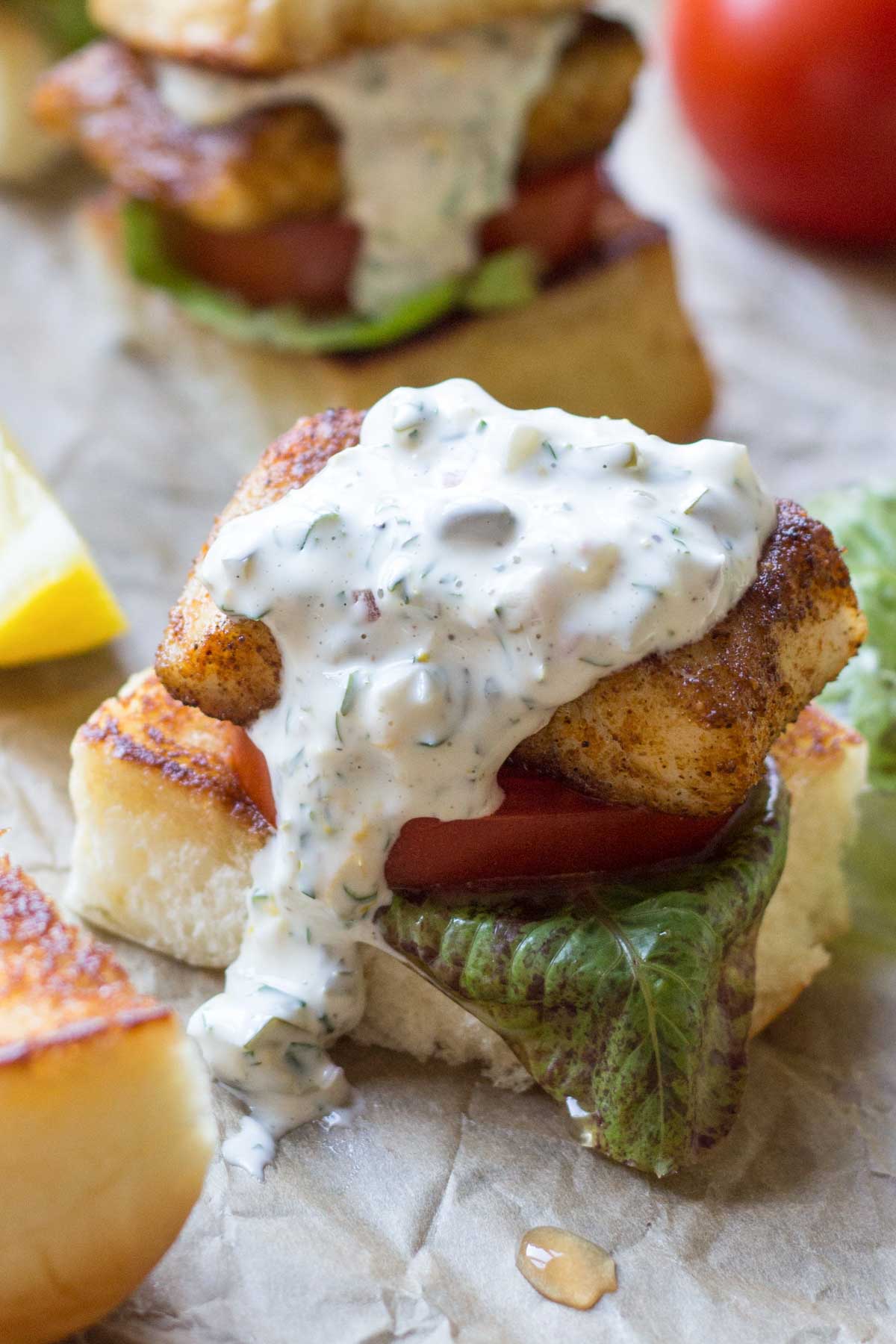 A piece of blackened fish on a toasted bun with lettuce and tomato, generously topped with creamy tartar sauce, which drips slightly down the side.