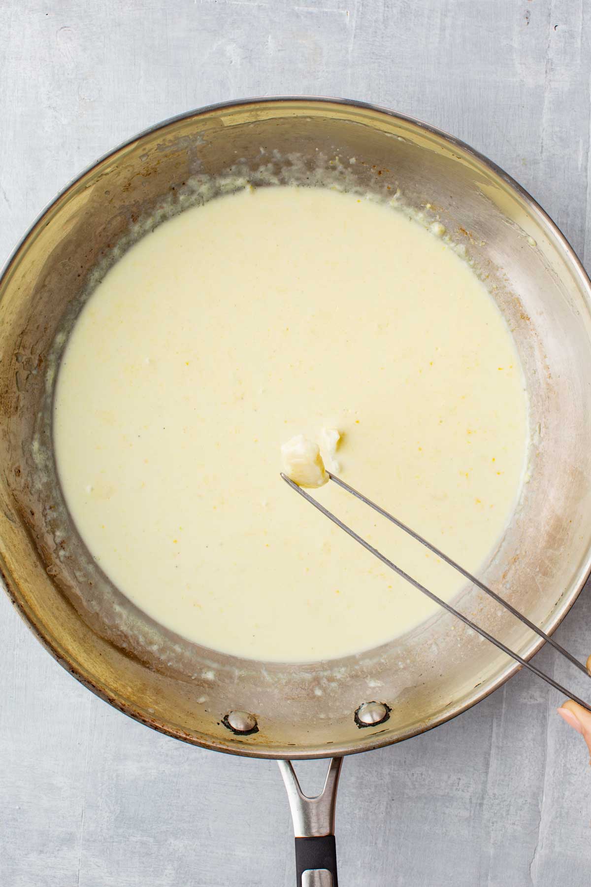 A creamy lemon sauce simmering in a skillet, with a hand holding tongs to remove a garlic clove after infusing the sauce.