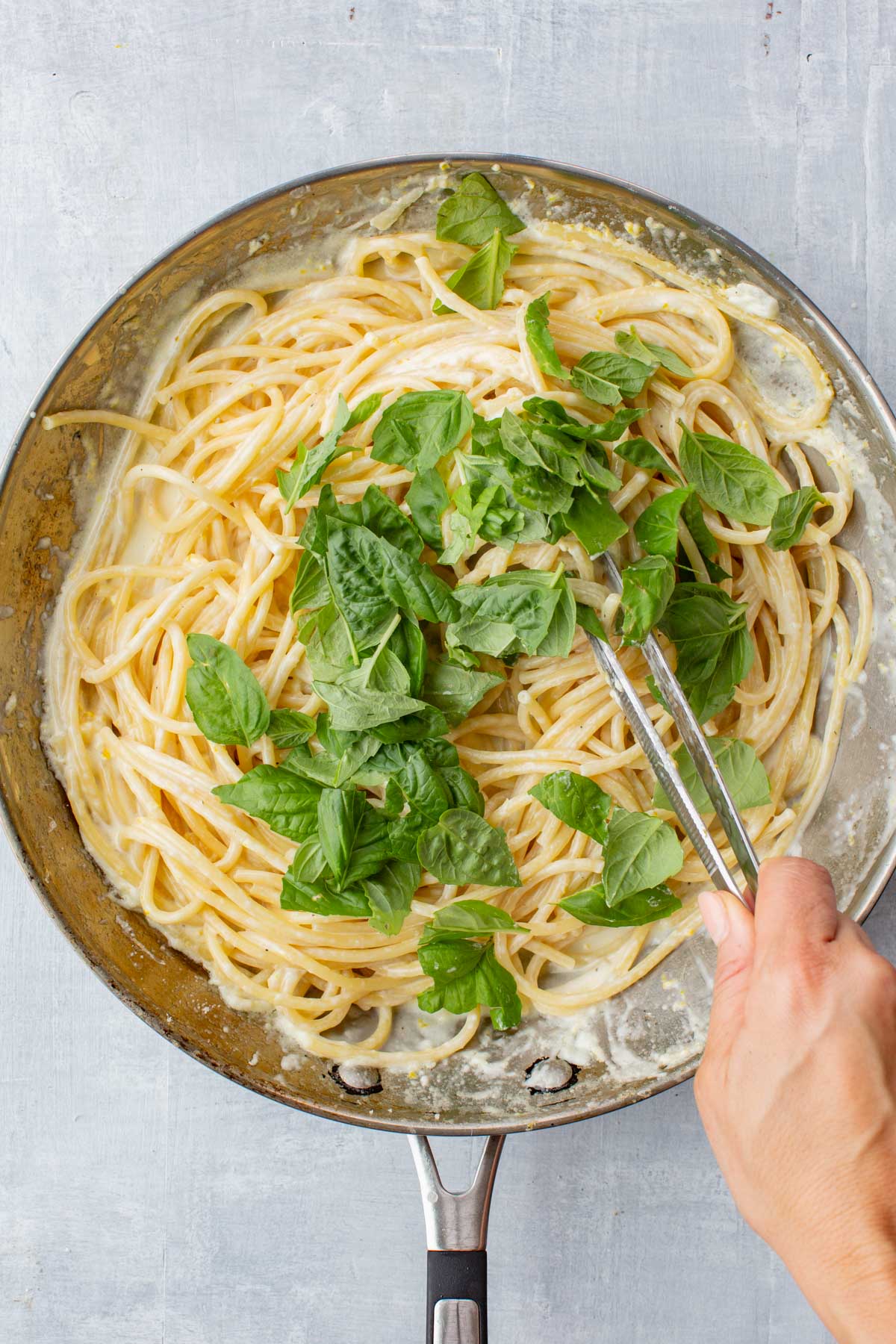 A hand using tongs to stir torn fresh basil into the creamy lemon spaghetti, adding a pop of green to the dish.