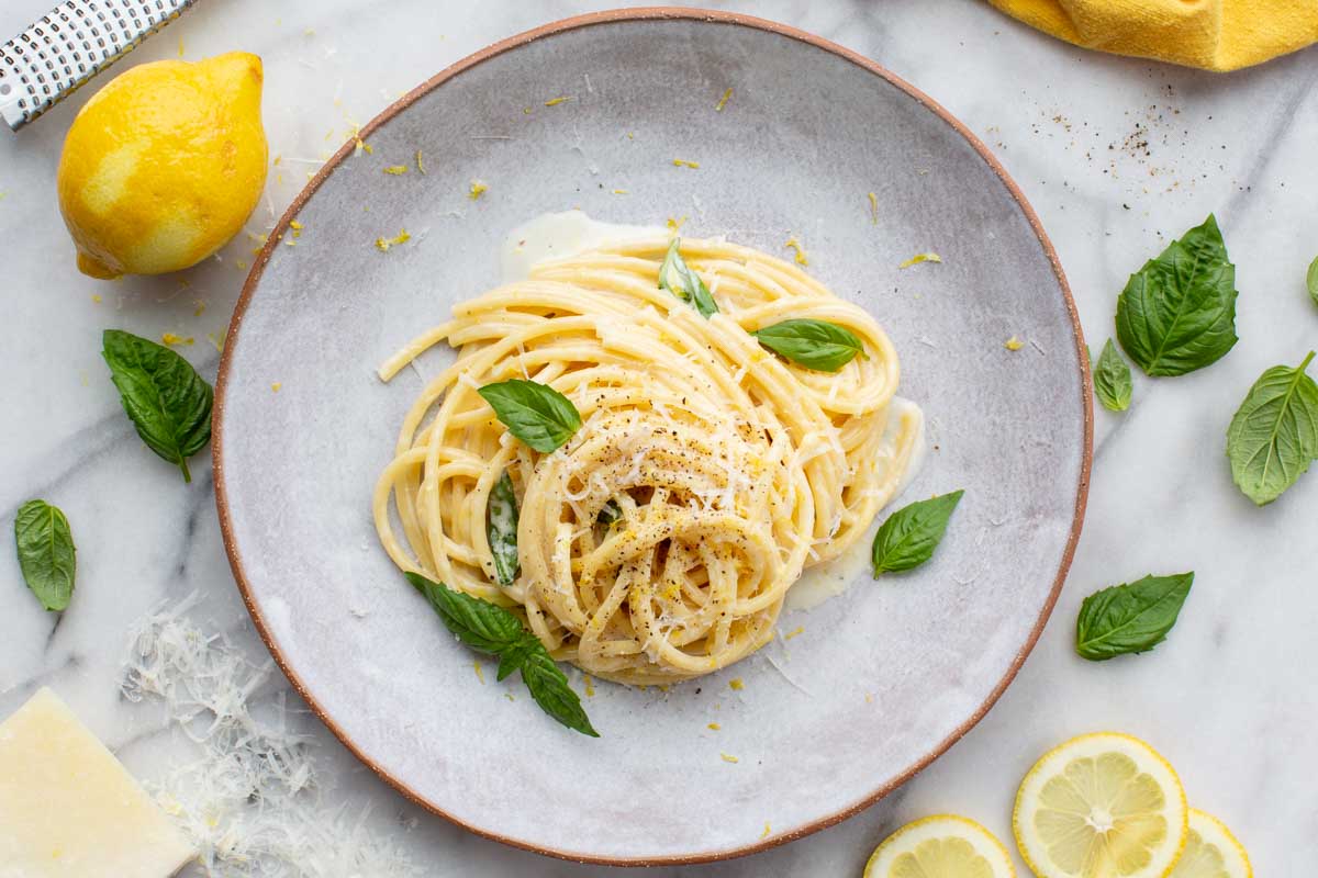 A plated serving of pasta al limone garnished with basil, black pepper, lemon zest, and surrounded by fresh lemon slices and grated cheese.