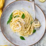 Top-down view of creamy pasta al limone on a ceramic plate with a fork and scattered basil leaves, Parmesan, and lemon rounds.