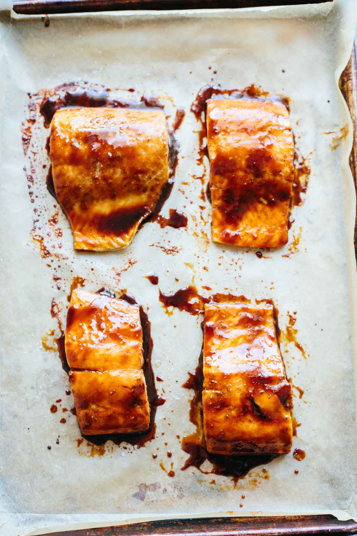 Four fillets of maple soy glazed salmon resting on a parchment-lined baking sheet, their shiny, caramelized surfaces glistening with sticky glaze.