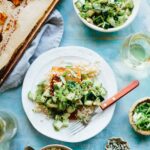 Overhead view of a bright blue table spread with cucumber avocado salad, white wine, a pan of baked salmon, and a plated salmon fillet served over rice.