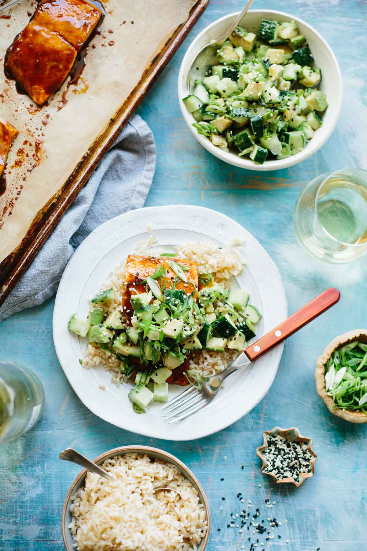 Overhead view of a bright blue table spread with cucumber avocado salad, white wine, a pan of baked salmon, and a plated salmon fillet served over rice.