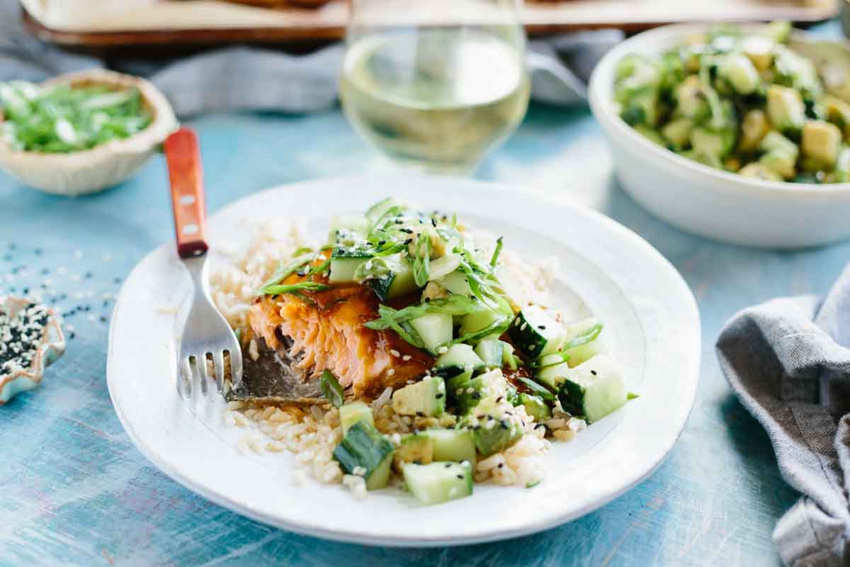 Close-up of a flaked salmon fillet revealing tender pink flesh underneath the glaze, topped with a crunchy cucumber avocado salad on a bed of rice.