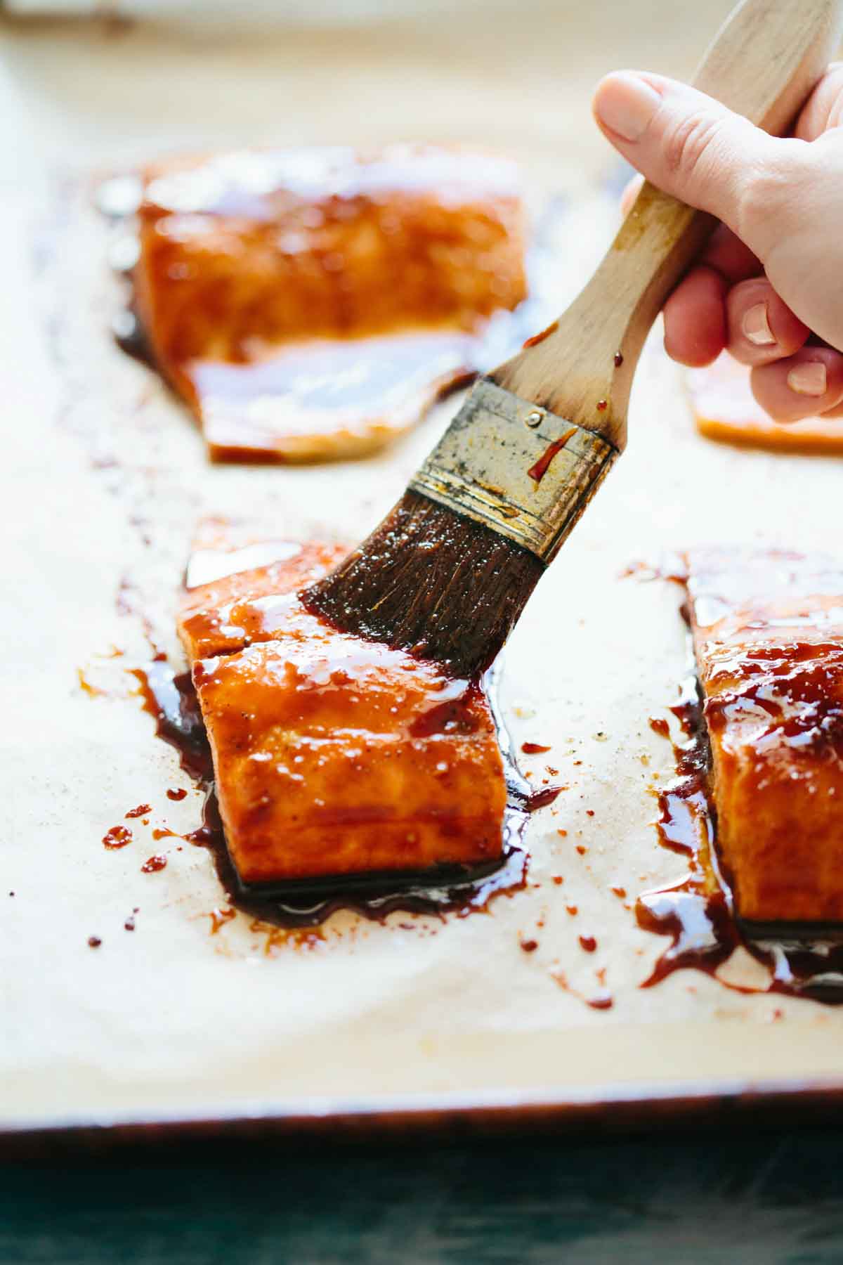A hand brushes rich, sticky maple soy glaze onto a piece of salmon with a pastry brush, set on a parchment-lined sheet pan.