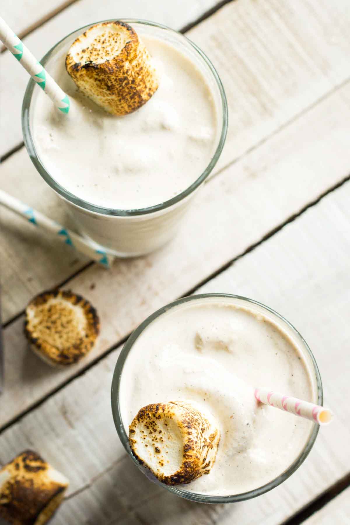 Two thick toasted marshmallow milkshakes served in tall glasses, topped with golden-brown marshmallows and colorful striped paper straws on a rustic whitewashed wooden table.