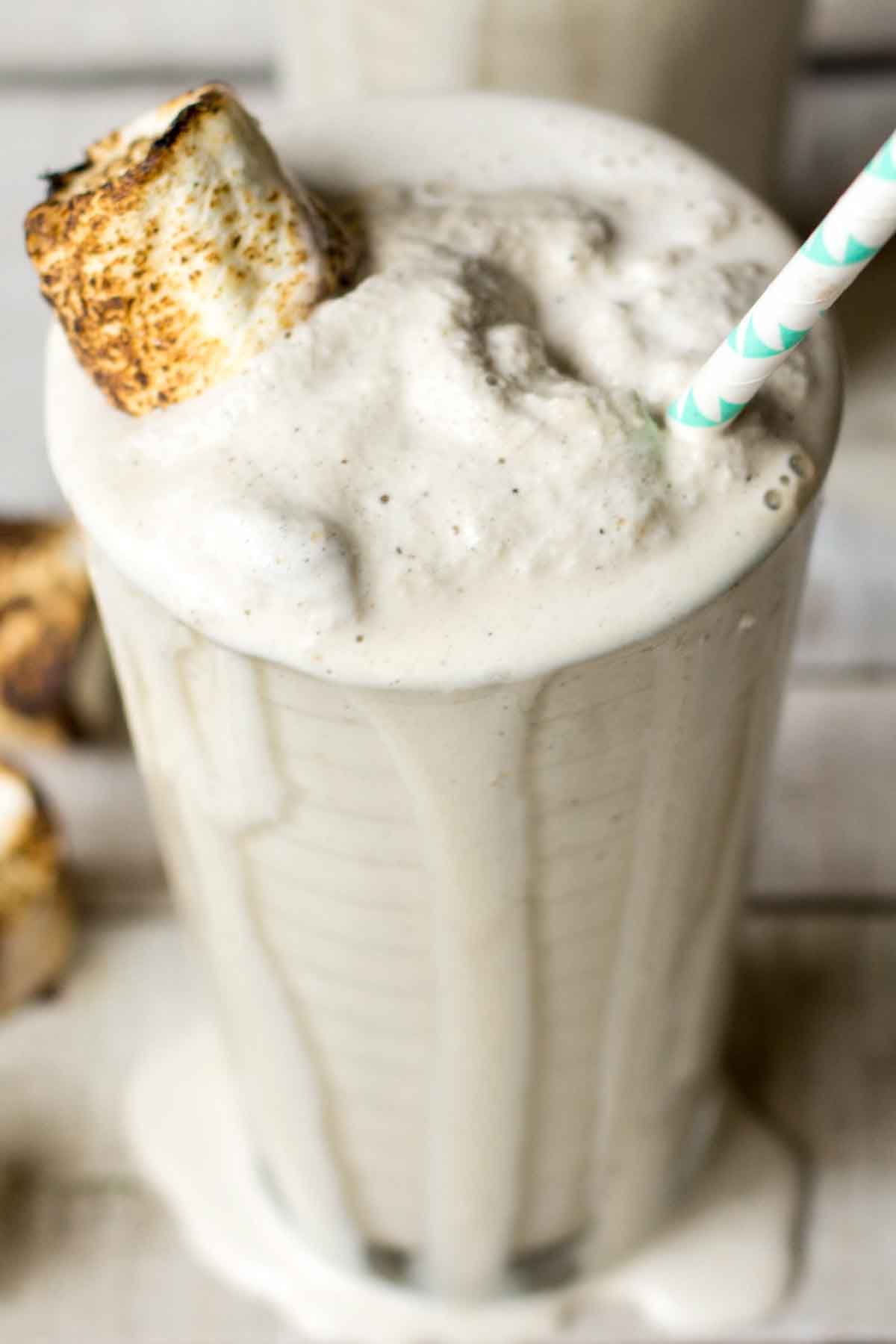 Close-up of a creamy toasted marshmallow milkshake overflowing slightly from a glass, with a toasted marshmallow and teal-striped straw nestled on top.