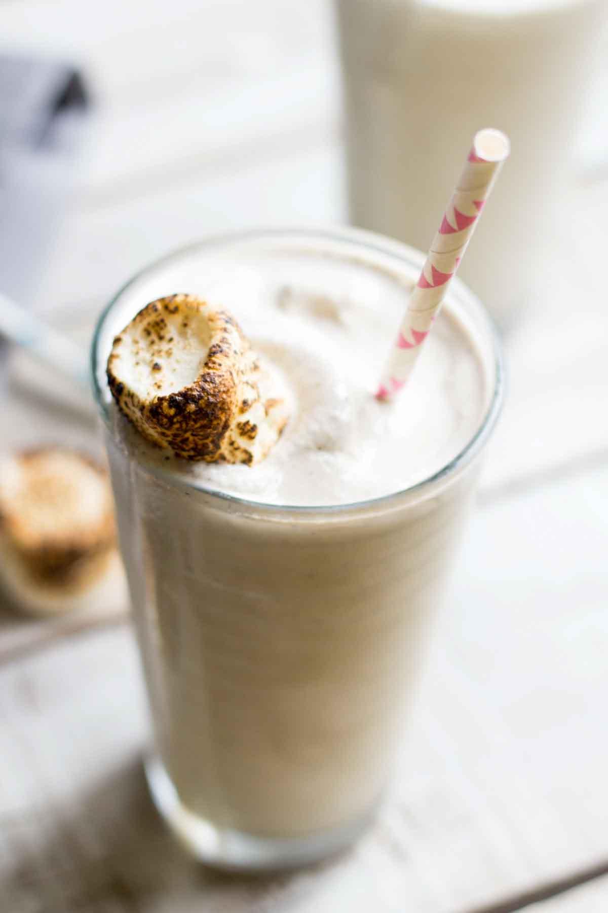 Side view of a toasted marshmallow milkshake in a tall clear glass with a pink-striped straw and a perfectly browned marshmallow garnish.