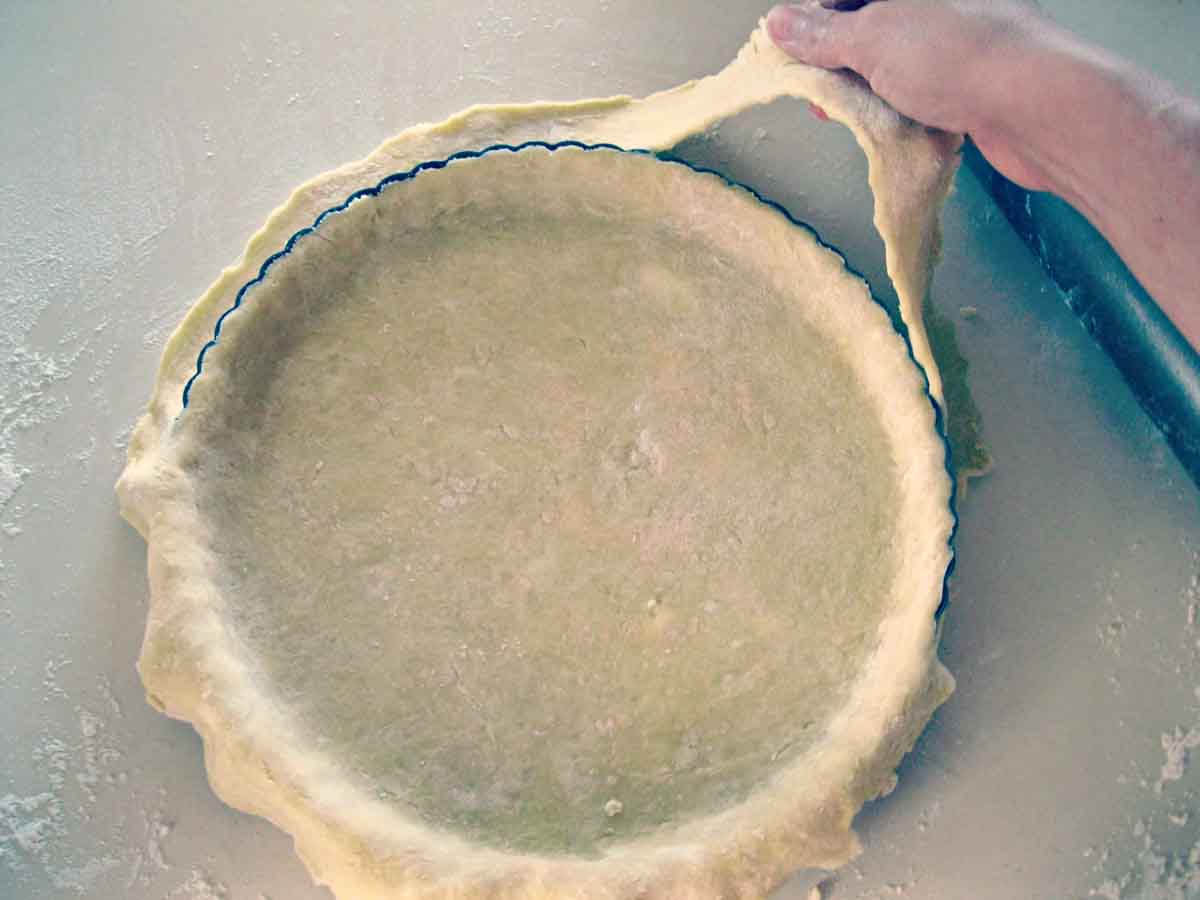 A hand carefully presses rolled-out tart dough into a fluted tart pan on a floured surface, preparing the crust for a savory tomato tart.