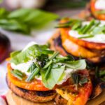 Close-up of a grilled toast layered with thick heirloom tomato slices, a dollop of creamy cheese, and a generous scatter of fresh basil, chives, and oregano on a wooden board.