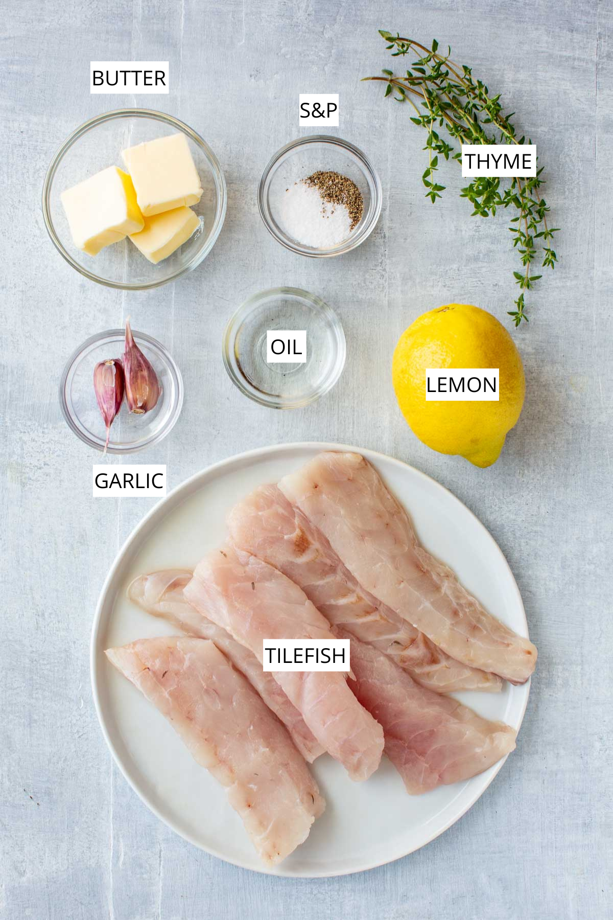 Overhead view of labeled ingredients for pan seared tilefish, including raw tilefish fillets on a white plate, a whole lemon, sprigs of fresh thyme, garlic cloves, butter cubes, neutral oil in a small bowl, and a dish of salt and black pepper.