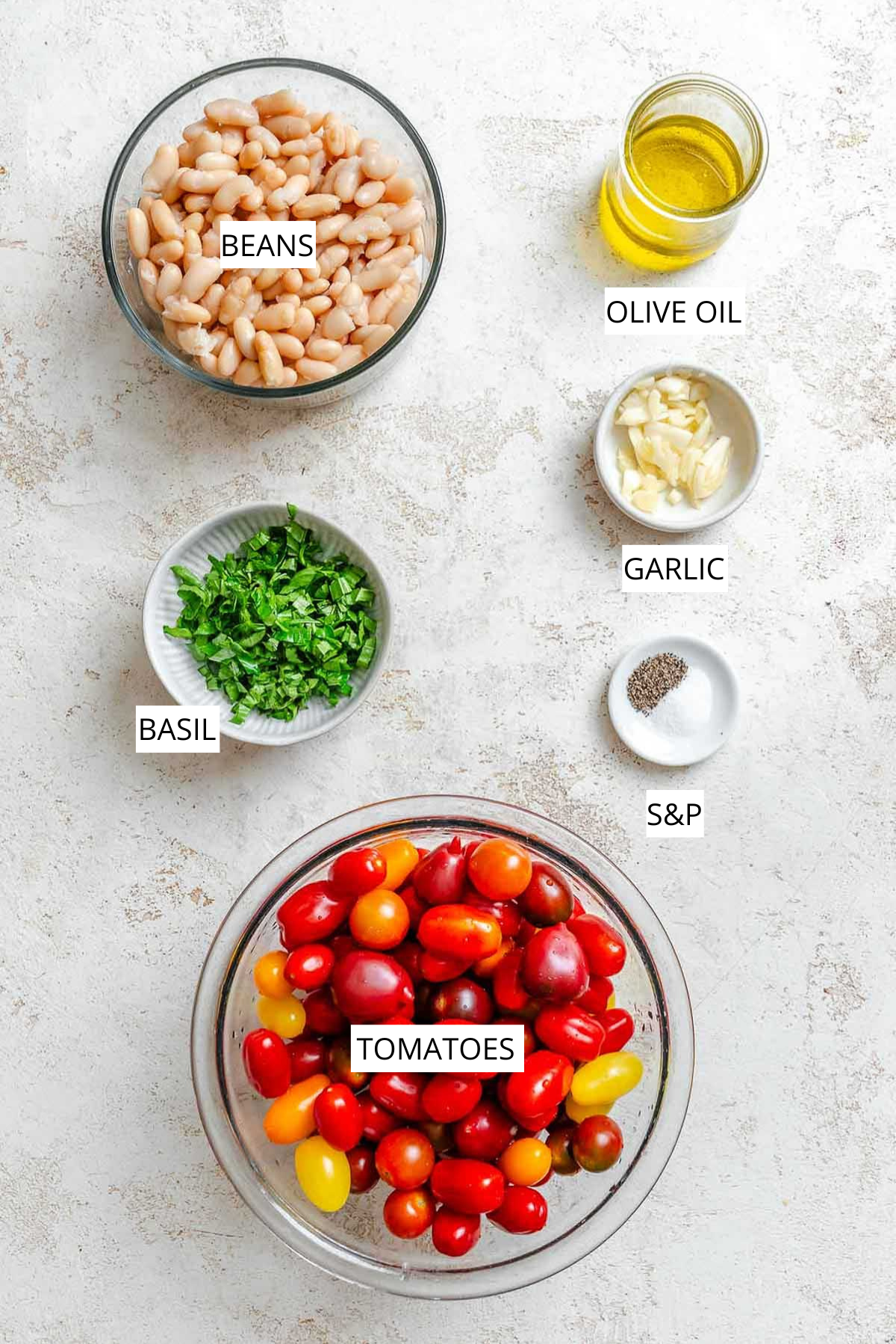 Overhead view of recipe ingredients in bowls: a bowl of white beans, a small jar of olive oil, sliced garlic cloves, chopped fresh basil, a dish of salt and black pepper, and a large bowl of colorful cherry tomatoes.