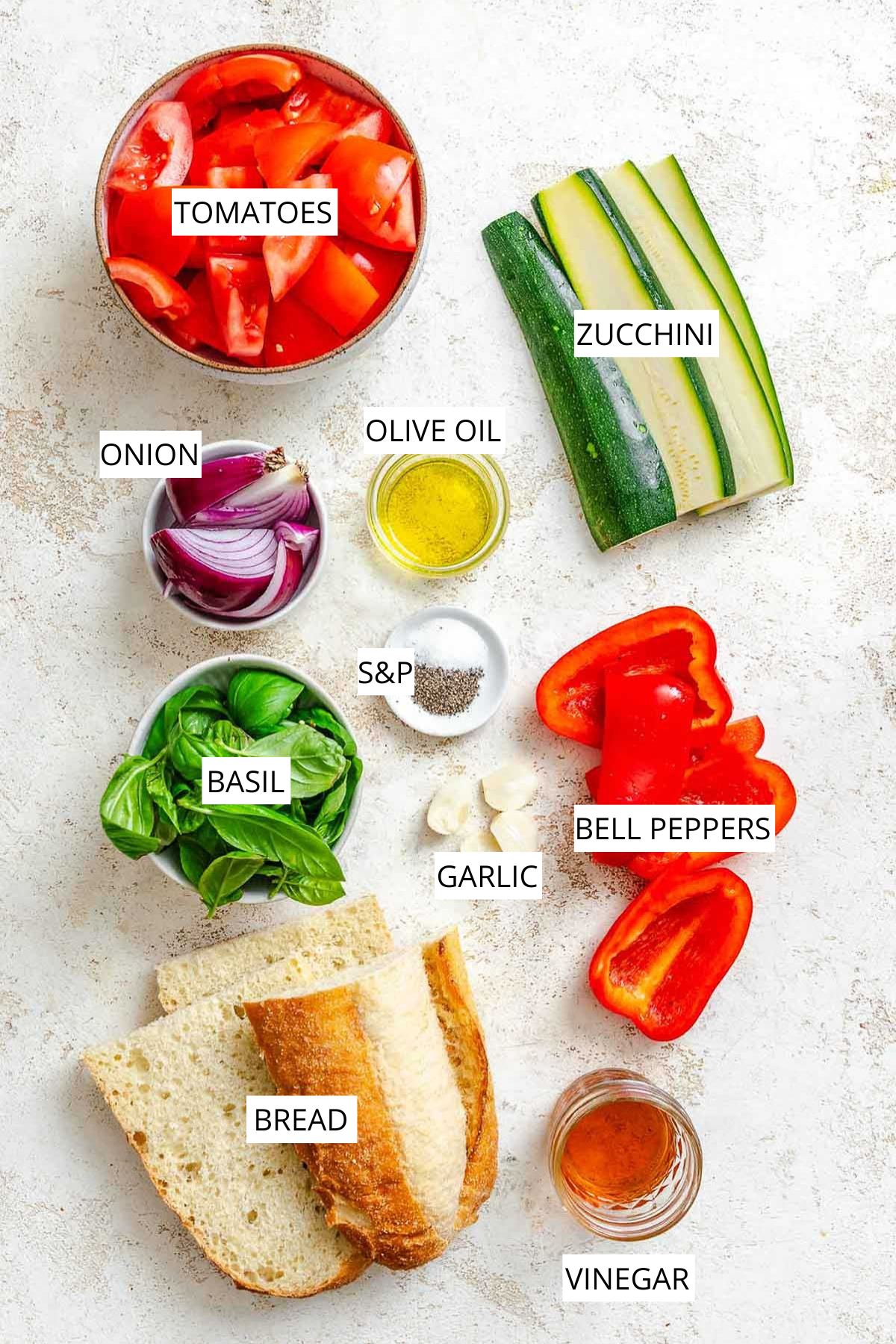 Overhead shot of ingredients labeled: tomatoes, zucchini, red onion, bell peppers, basil, garlic, olive oil, vinegar, bread, salt, and pepper.