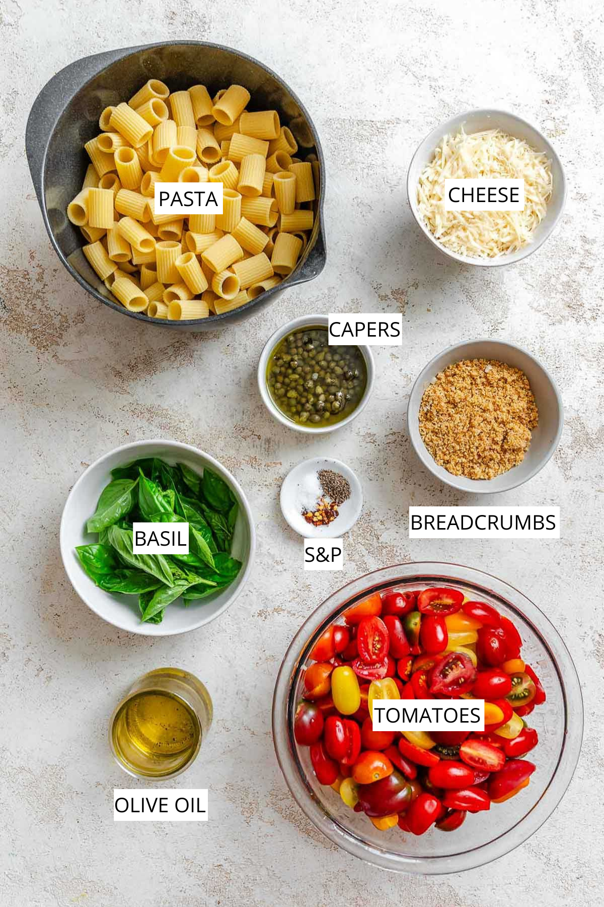 Overhead view of labeled Pasta Ponza ingredients on a light surface, including a bowl of rigatoni pasta, shredded cheese, capers in brine, toasted breadcrumbs, fresh basil leaves, salt and pepper with red pepper flakes, olive oil, and halved cherry and grape tomatoes in various colors.