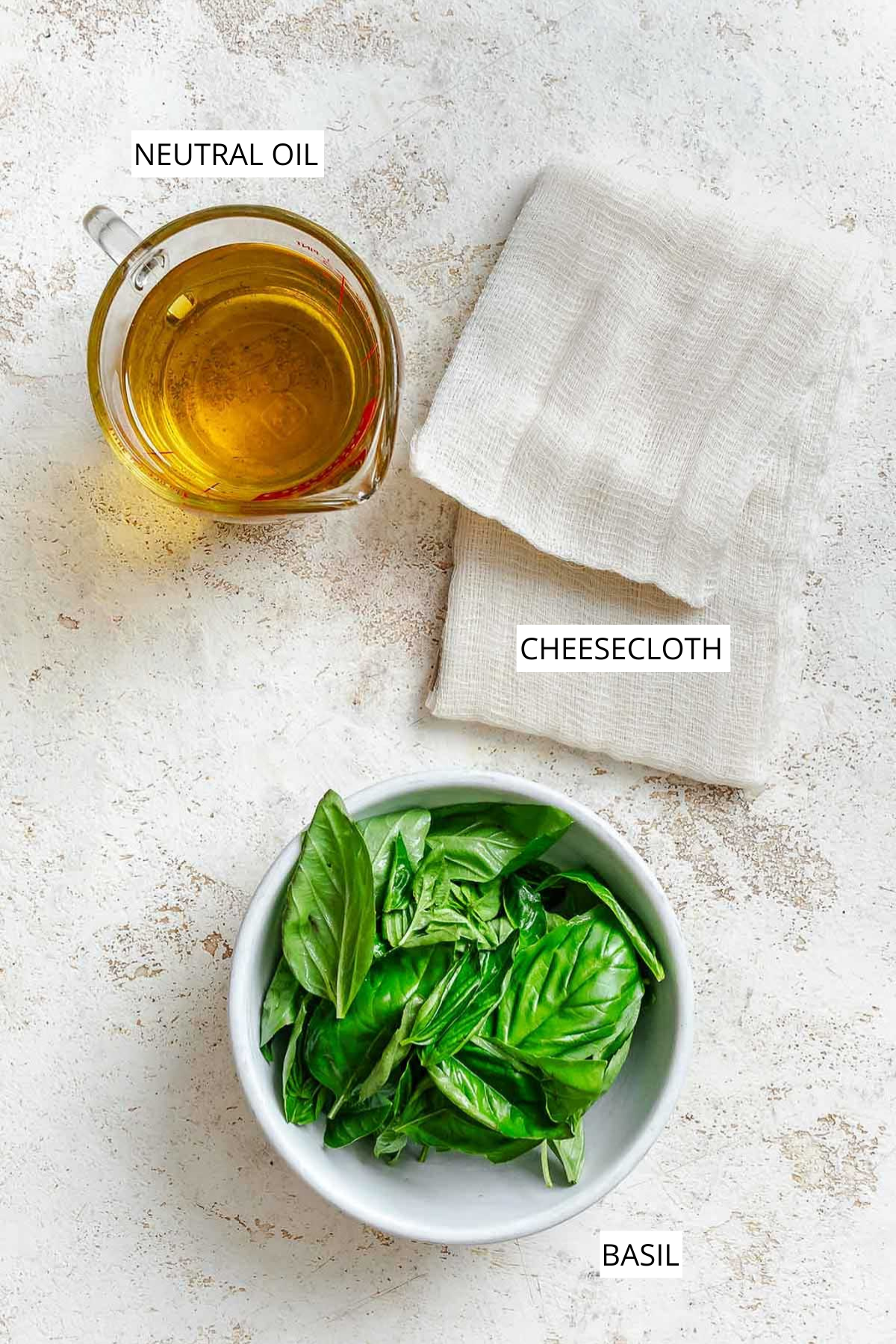 Overhead view of measured ingredients for homemade basil oil, including a glass measuring cup of neutral oil, folded cheesecloth, and a white bowl filled with fresh basil leaves, arranged on a light textured surface.