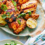 Platter of grilled chicken thighs with charred lime - Golden, smoky chicken thighs garnished with fresh cilantro sprigs and halved limes with grill marks, ready to be served.