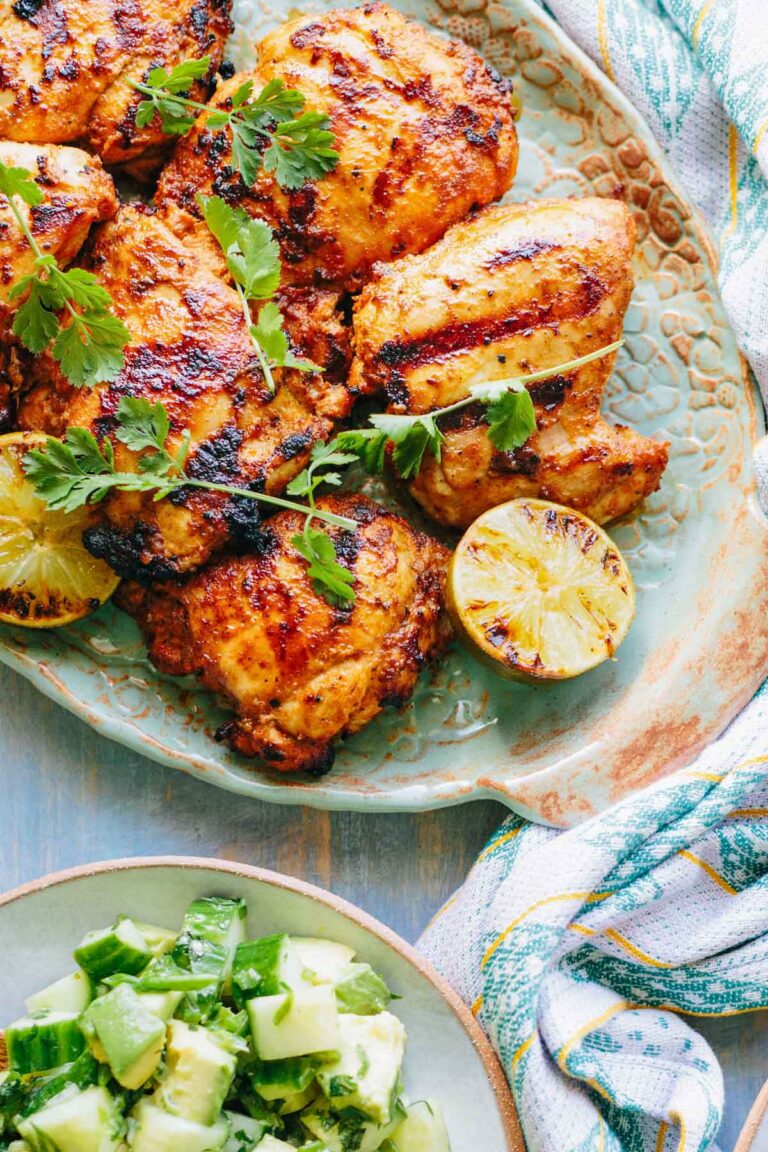 Platter of grilled chicken thighs with charred lime – Golden, smoky chicken thighs garnished with fresh cilantro sprigs and halved limes with grill marks, ready to be served.