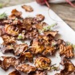 Close-up of beet chips with fresh beets - Golden and crimson beet chips on parchment paper with raw beets and leafy tops blurred in the background, sprinkled with herbs and flaky salt.