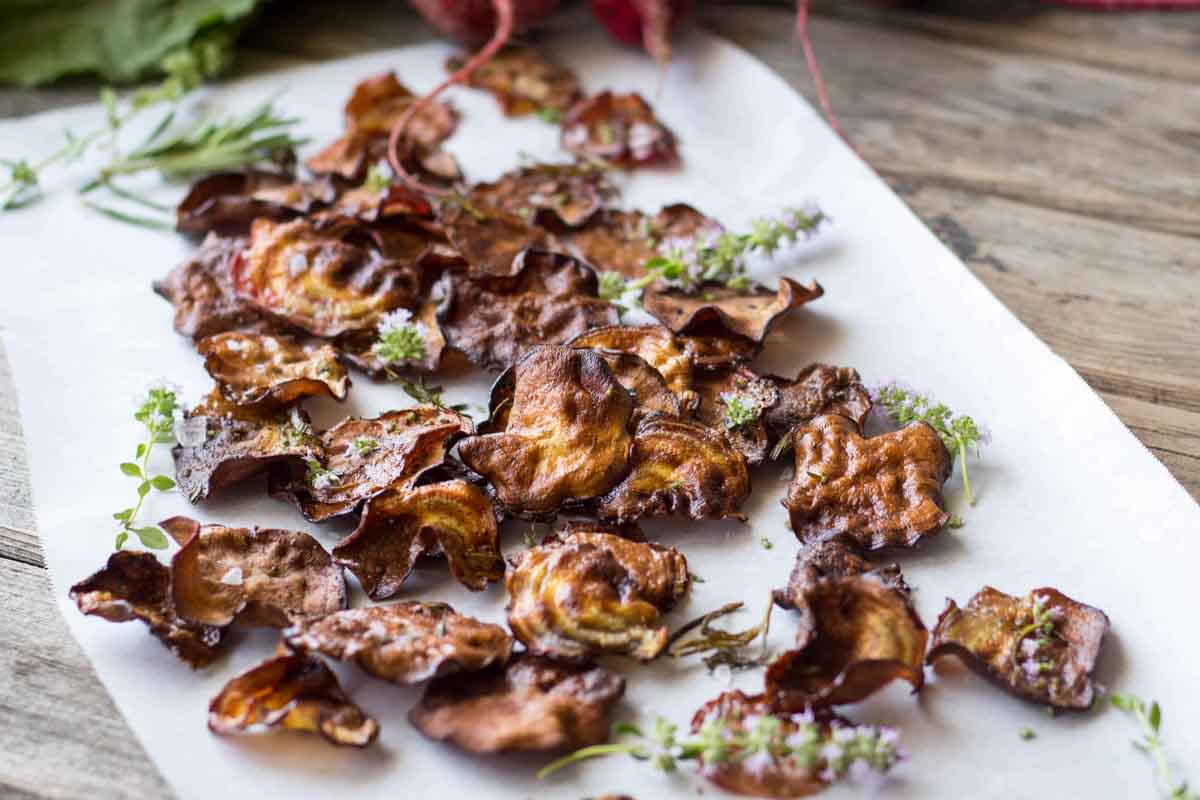 Rustic beet chips on parchment - Crispy oven-roasted beet chips scattered over parchment paper and garnished with fresh herbs on a wooden surface.