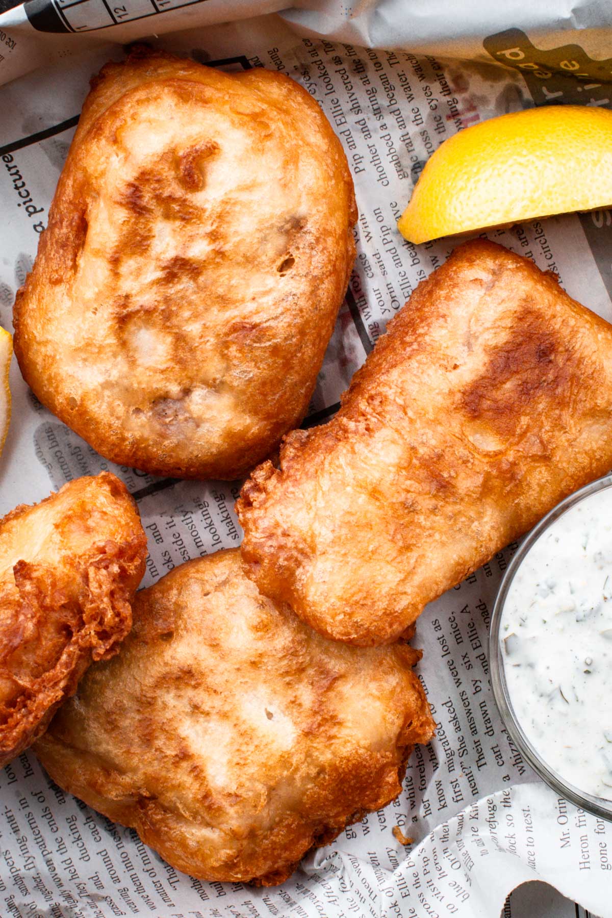 Crispy beer-battered fish on newspaper with lemon wedges and a small bowl of tartar sauce.