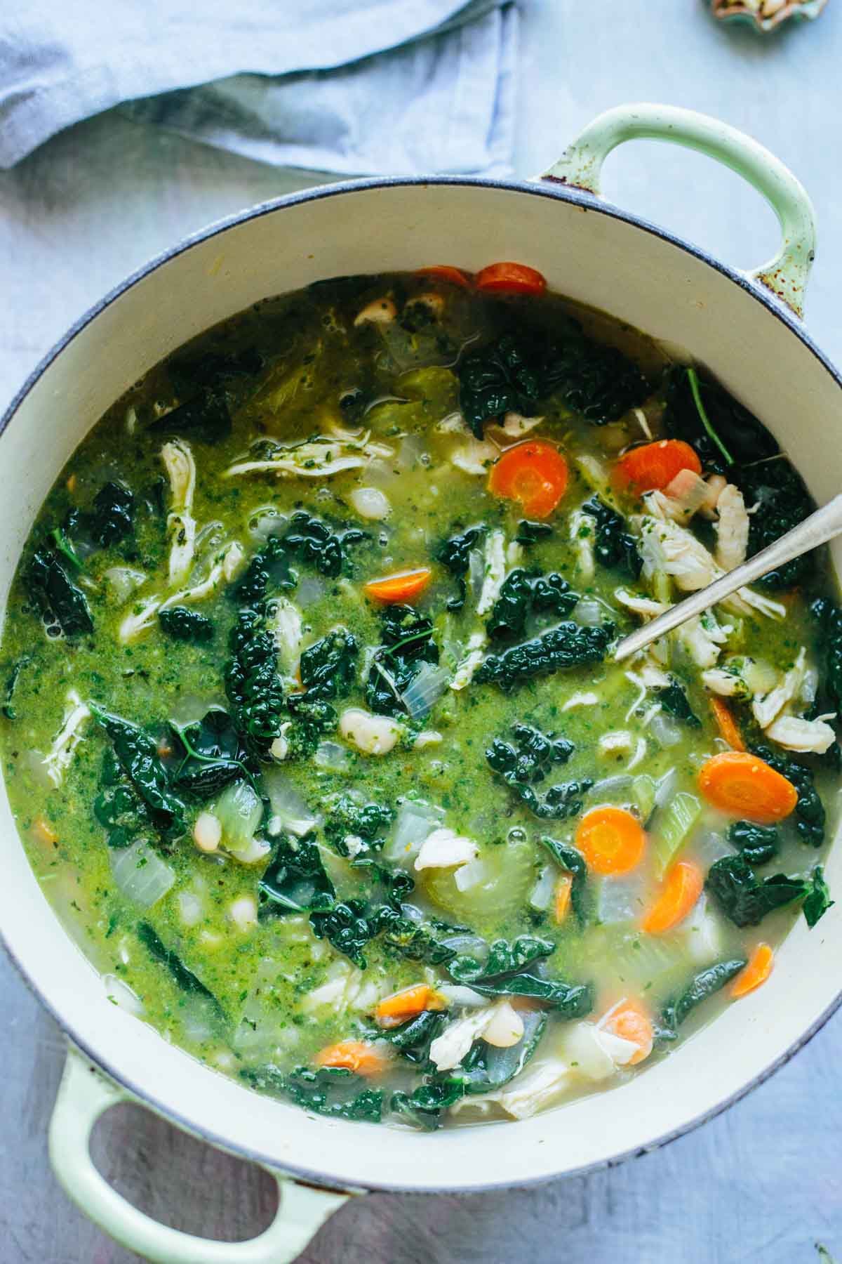 Overhead view of a large enamel pot filled with chicken soup with pesto, kale, white beans, carrots, and onions simmering in broth.