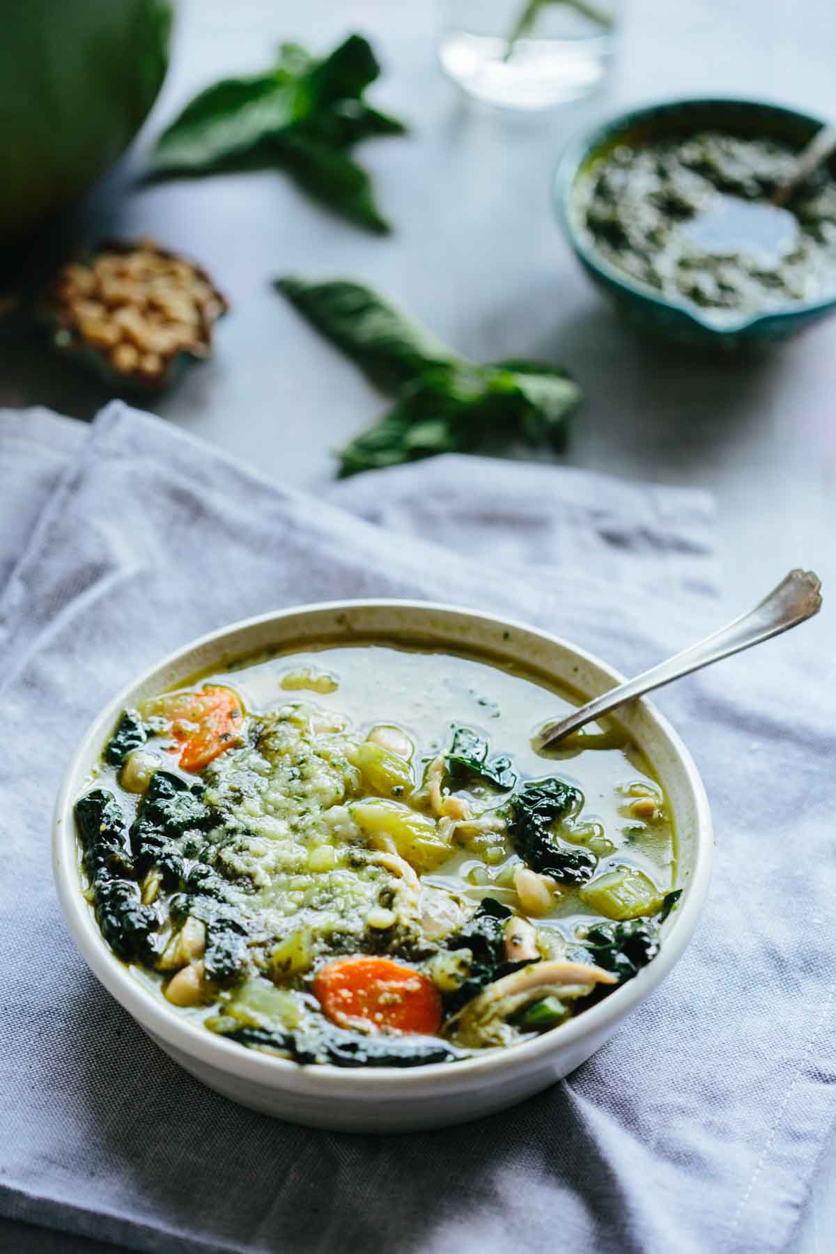 Angled shot of the soup bowl with basil leaves and a small dish of pesto in the background for garnish context.