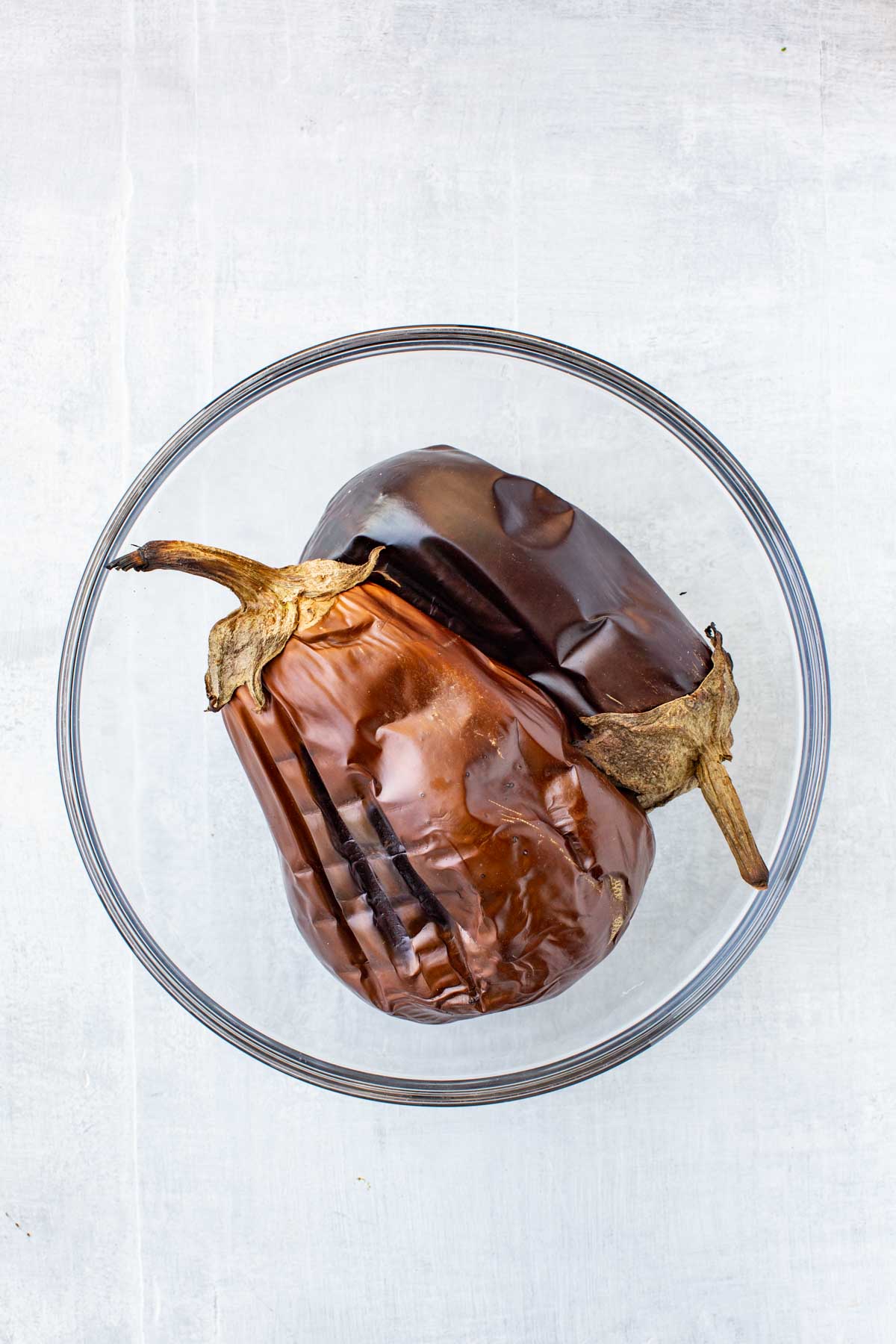 Whole charred eggplants in a glass bowl - Two roasted whole eggplants with wrinkled, blackened skin resting in a clear bowl after being charred.