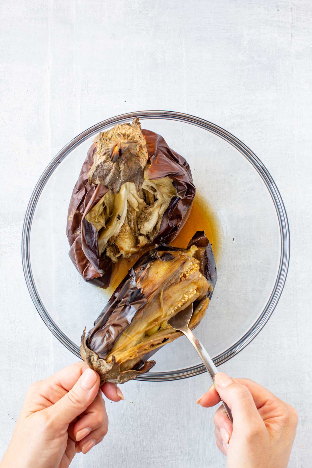 Peeling roasted eggplant - Hands using a spoon to peel open a roasted eggplant in a glass bowl, revealing the soft, golden flesh inside.