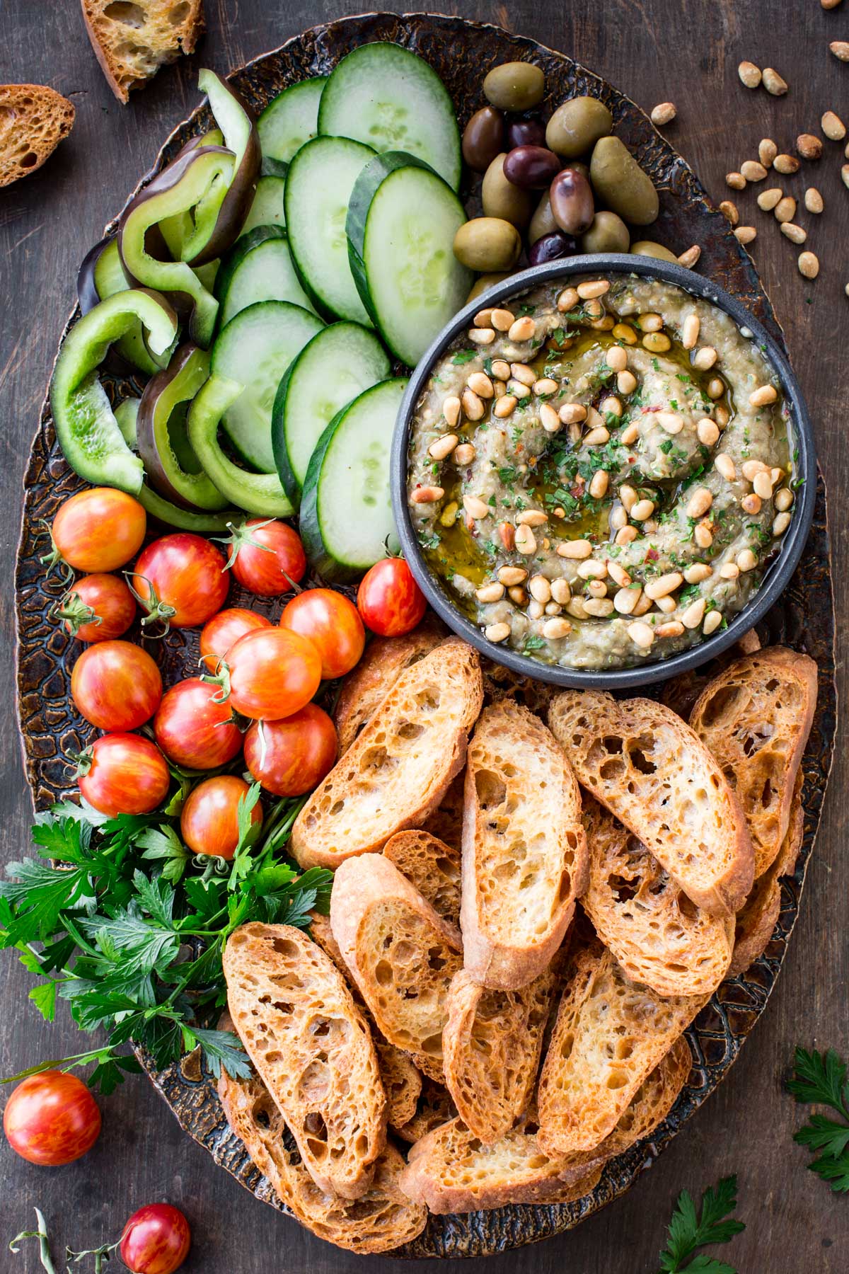 Eggplant dip platter with accompaniments - A serving platter filled with charred eggplant dip topped with pine nuts, surrounded by crusty bread slices, cucumber slices, cherry tomatoes, green peppers, olives, and fresh parsley.