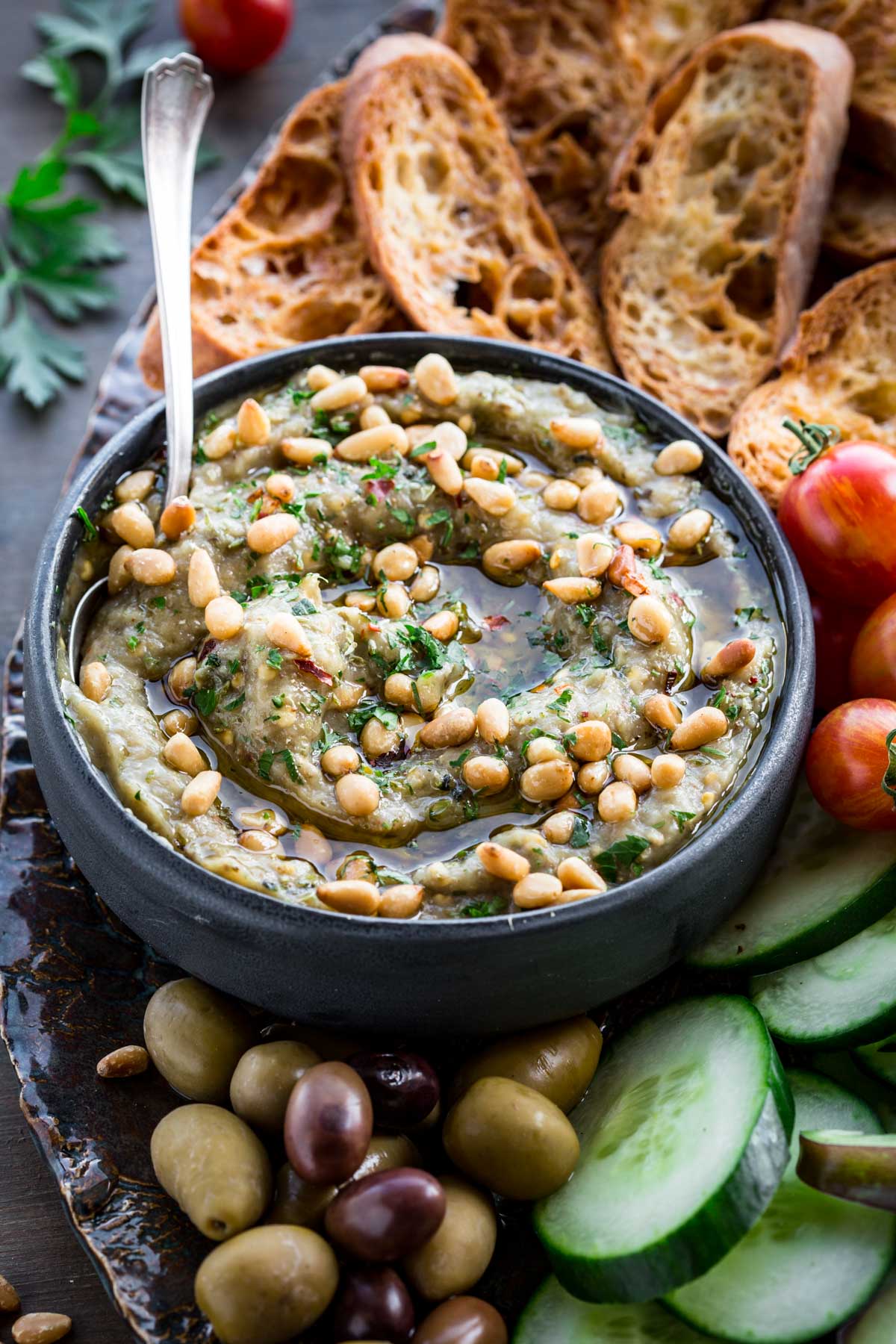 Eggplant dip with bread and vegetables - A black bowl of smoky eggplant dip with pine nuts, surrounded by cucumber slices, cherry tomatoes, olives, and crusty bread on a serving board.