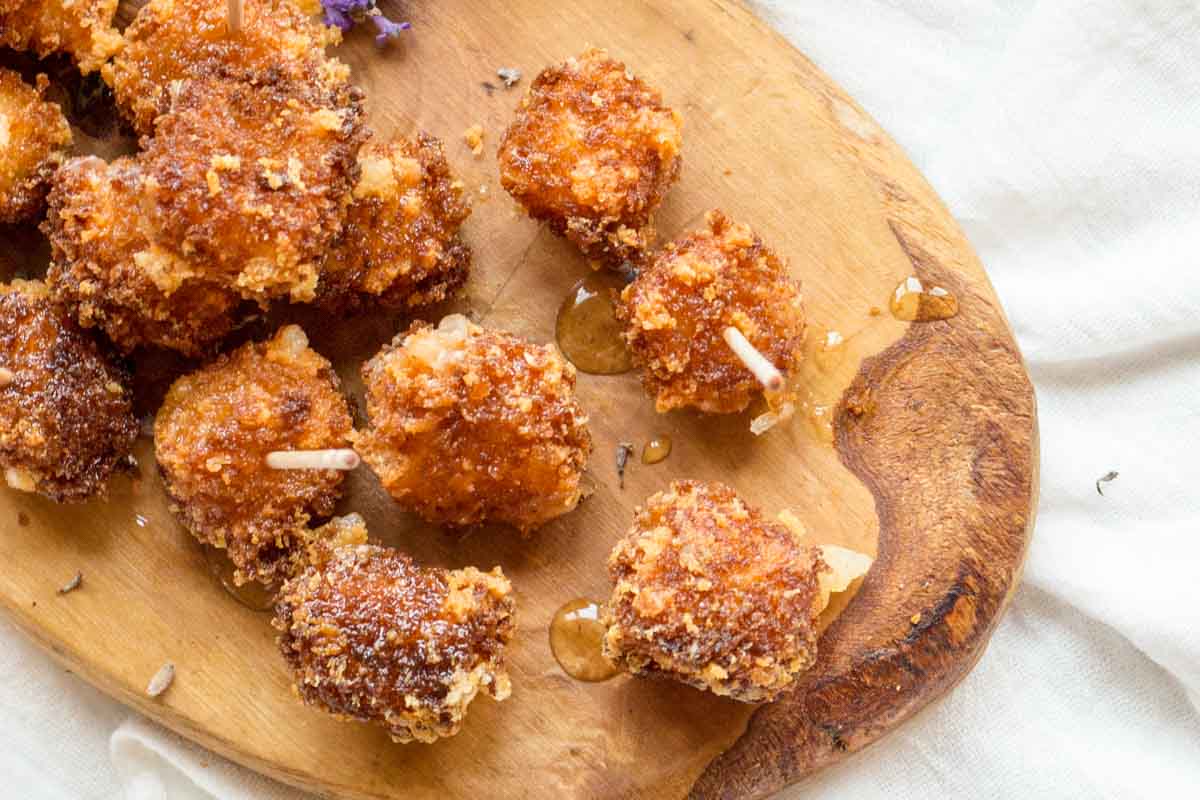 Overhead view of cheese bites and honey - Crunchy Manchego fritters scattered across a rustic wood board, glistening with honey and garnished with dried lavender.